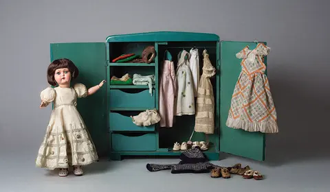 A 1930s composition doll in a cream lace dress standing beside its open wooden wardrobe trunk