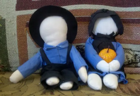 Two faceless Amish cloth dolls, a boy and a girl in traditional dress, seated together