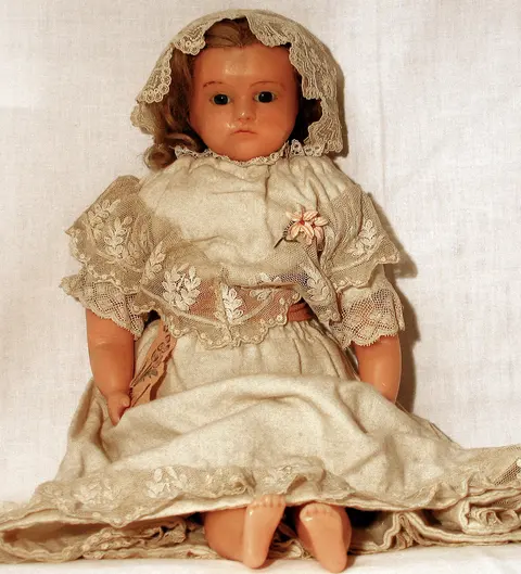 An antique wax doll wearing a lace bonnet and cream lace-trimmed Victorian dress, seated